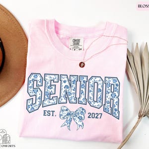 a pink t-shirt with the text "SENIOR" and "EST. 2027" printed on it, along with a bow design. The t-shirt is laid out on a white surface, accompanied by a brown hat, a necklace, and a palm frond.