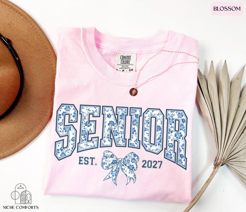 a pink t-shirt with the text "SENIOR" and "EST. 2027" printed on it, along with a bow design. The t-shirt is laid out on a white surface, accompanied by a brown hat, a necklace, and a palm frond.