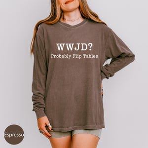 A woman wearing a brown long-sleeved shirt with the text "WWJD?" printed on it, posing against a plain background.
