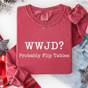 A red t-shirt with the text "WWJD?" and "Probably Flip Tables" printed on it, along with a necklace and a pair of jeans, all laid out on a white background.