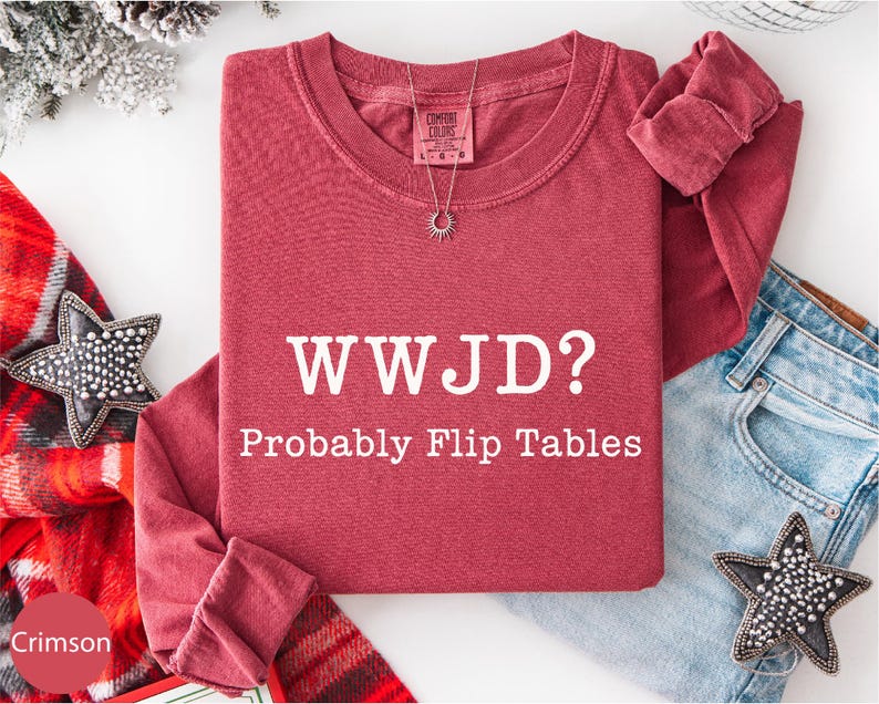 A red t-shirt with the text "WWJD?" and "Probably Flip Tables" printed on it, along with a necklace and a pair of jeans, all laid out on a white background.