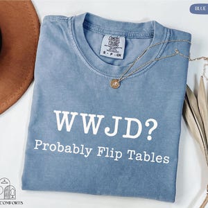 A blue t-shirt with the text "WWJD?" printed on it, along with the phrase "Probably Flip Tables" below it. The t-shirt is laid out on a white surface, with a brown hat and some dried leaves visible in the background.