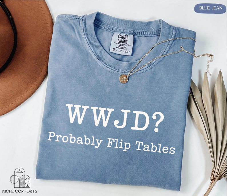 A blue t-shirt with the text "WWJD?" printed on it, along with the phrase "Probably Flip Tables" below it. The t-shirt is laid out on a white surface, with a brown hat and some dried leaves visible in the background.
