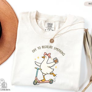 a white t-shirt with a cartoon image of a duck riding a scooter and holding a coffee cup, accompanied by the text "Off to regulate emotions".