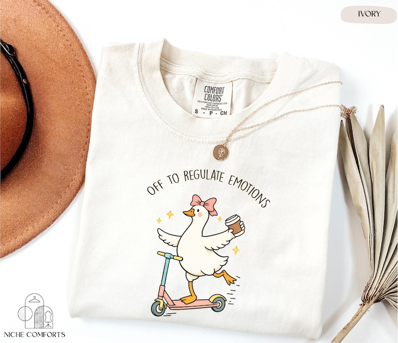 a white t-shirt with a cartoon image of a duck riding a scooter and holding a coffee cup, accompanied by the text "Off to regulate emotions".