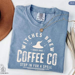 May include: A blue jean colored t-shirt with the text "Witches Brew Coffee Co" printed in white. The shirt features a round neck and short sleeves. A gold necklace with a pendant is draped across the shirt.