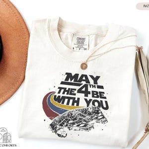 May include: Ivory-colored t-shirt with the text "May the 4th Be With You" and a graphic of a spaceship. The shirt has a crew neck and short sleeves. A gold necklace with a pendant is draped on the shirt.