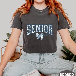 A woman wearing a black t-shirt with the text "SENIOR EST. 2027" printed on it, sitting on a bench with a potted plant in the background.