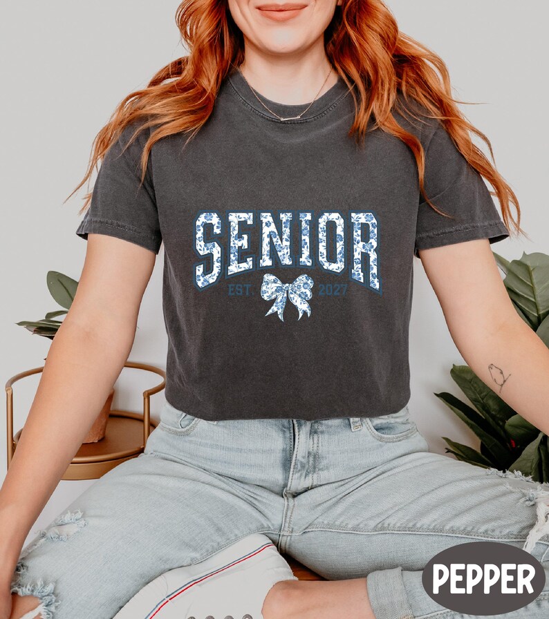 A woman wearing a black t-shirt with the text "SENIOR EST. 2027" printed on it, sitting on a bench with a potted plant in the background.
