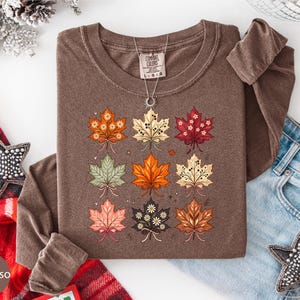May include: A long-sleeve brown t-shirt featuring a graphic print of nine stylized maple leaves arranged in a grid. Each leaf displays a unique color palette, including orange, cream, red, green, and dark brown, with small floral and dot accents.