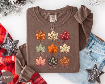 May include: A long-sleeve brown t-shirt featuring a graphic print of nine stylized maple leaves arranged in a grid. Each leaf displays a unique color palette, including orange, cream, red, green, and dark brown, with small floral and dot accents.