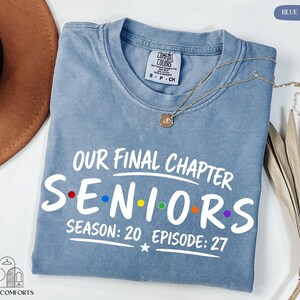 May include: A blue jean colored t-shirt with white text that reads Our Final Chapter Seniors Season: 20 Episode: 27. The word Seniors features small colored dots between the letters. The shirt has a crew neckline and a Comfort Colors brand label.