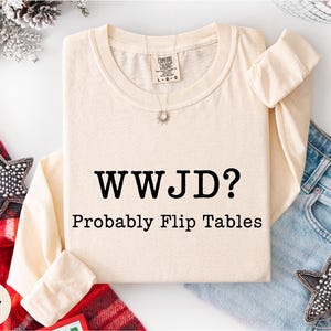 A beige long-sleeved shirt with the text "WWJD?" printed on it, along with the caption "Probably Flip Tables" below it. The shirt is laid out on a white background, with a pair of jeans and a red and white striped scarf nearby.