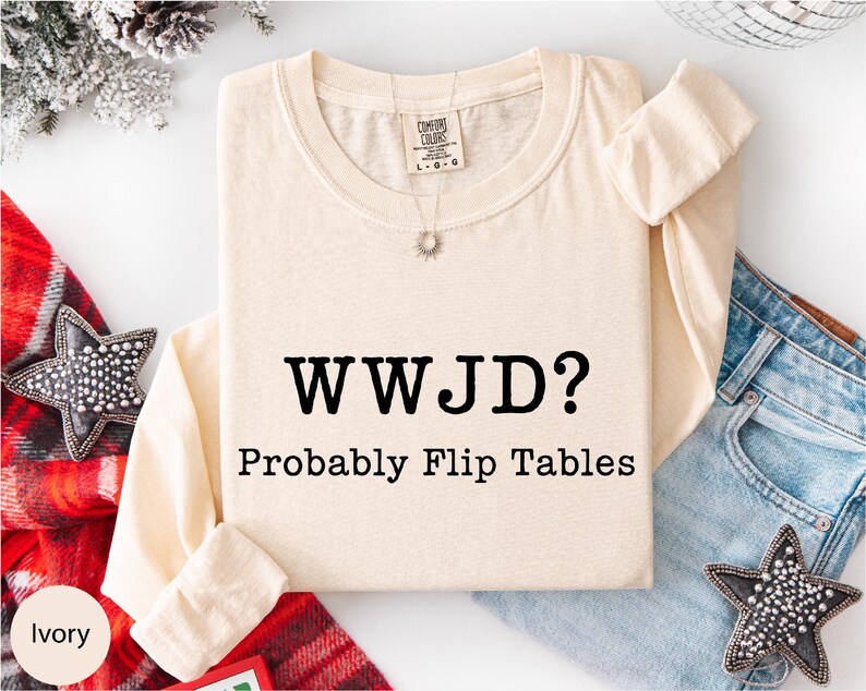 A beige long-sleeved shirt with the text "WWJD?" printed on it, along with the caption "Probably Flip Tables" below it. The shirt is laid out on a white background, with a pair of jeans and a red and white striped scarf nearby.