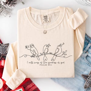 A beige long-sleeved t-shirt with a black line drawing of three birds and the text "I will sing of the goodness of God" printed on it, along with a necklace and a star-shaped embellishment.
