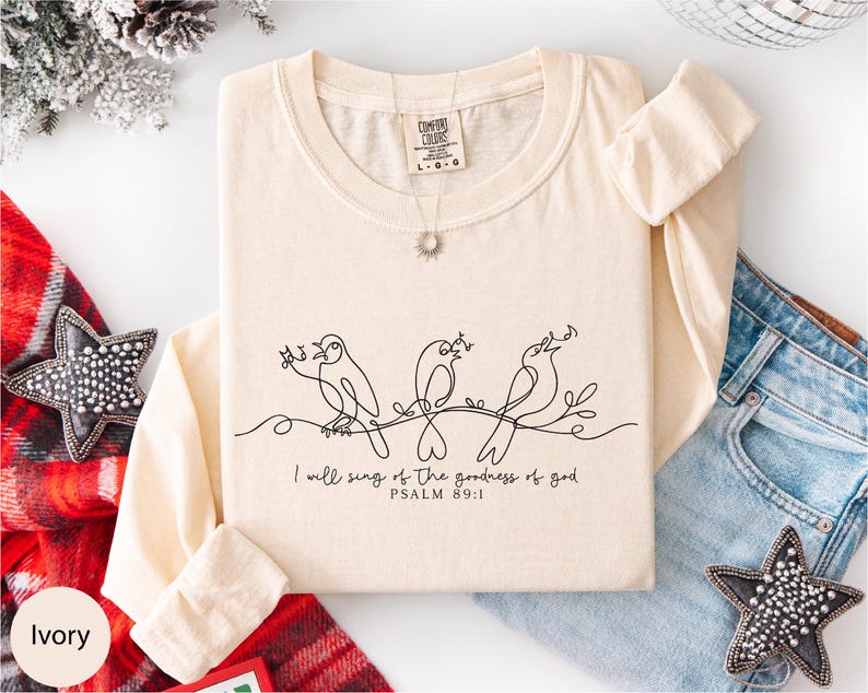A beige long-sleeved t-shirt with a black line drawing of three birds and the text "I will sing of the goodness of God" printed on it, along with a necklace and a star-shaped embellishment.