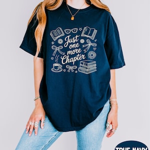 A woman wearing a navy blue t-shirt with the text "Just one more chapter" printed on it, along with various icons and symbols related to reading and writing.