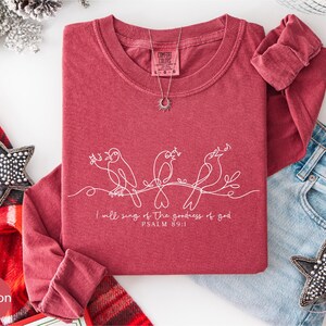 A red t-shirt with a white design of birds and text, along with a pair of jeans and a star-shaped ornament, are arranged on a white background.