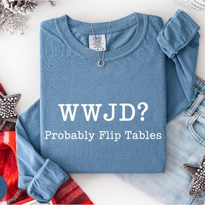 A blue t-shirt with the text "WWJD?" and "Probably Flip Tables" printed on it, along with a necklace and a pair of jeans, are displayed on a white surface.