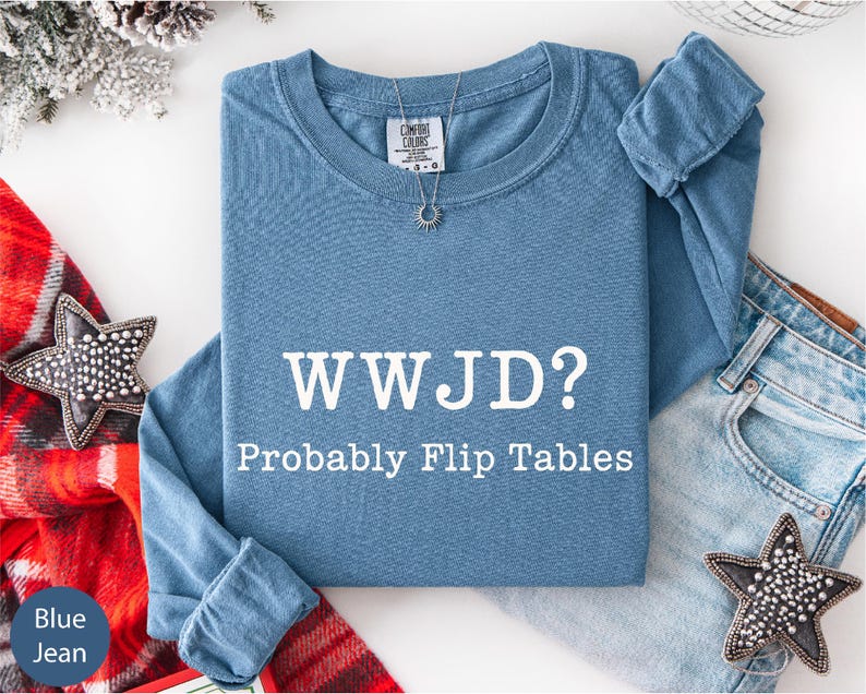 A blue t-shirt with the text "WWJD?" and "Probably Flip Tables" printed on it, along with a necklace and a pair of jeans, are displayed on a white surface.