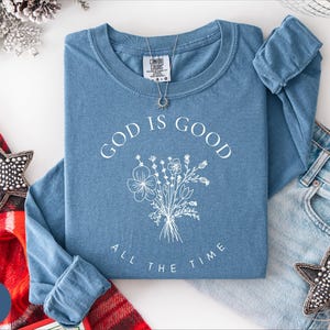 May include: A blue jean long-sleeve shirt with the text "GOD IS GOOD ALL THE TIME" and a white floral bouquet design. A silver necklace and a pair of blue jeans are next to the shirt. A red and black plaid blanket and a star-shaped ornament are also in the image.