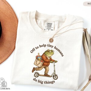 May include: Ivory-colored t-shirt with a graphic of a frog riding a scooter, carrying a bag of school supplies. Text reads "Off to help tiny humans do big things." The shirt has a crew neck and short sleeves.