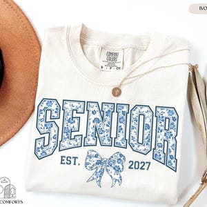 a white t-shirt with the text "SENIOR EST. 2027" printed on it, along with a blue floral design. The t-shirt is laid out on a white background, accompanied by a brown hat, a necklace, and some dried leaves.