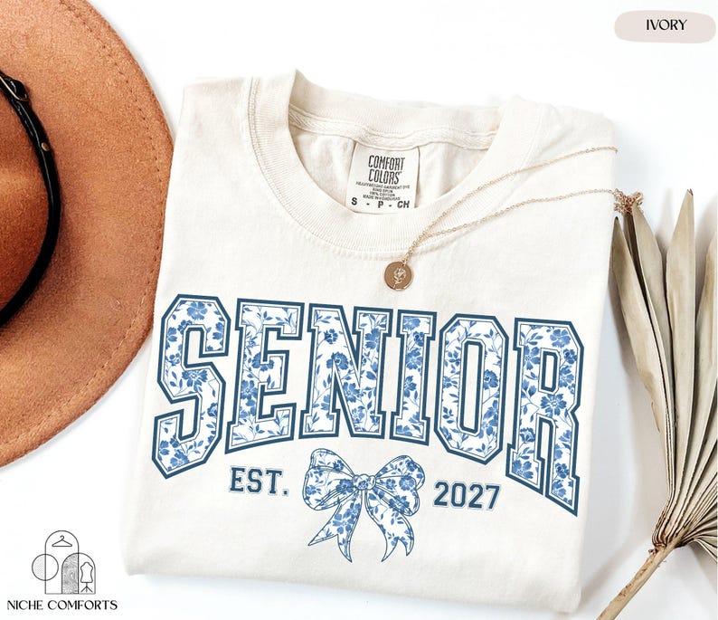 a white t-shirt with the text "SENIOR EST. 2027" printed on it, along with a blue floral design. The t-shirt is laid out on a white background, accompanied by a brown hat, a necklace, and some dried leaves.