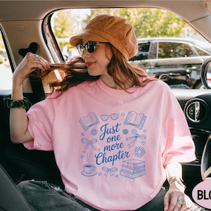 A woman wearing a pink t-shirt with a graphic design and text, sitting in the passenger seat of a car.