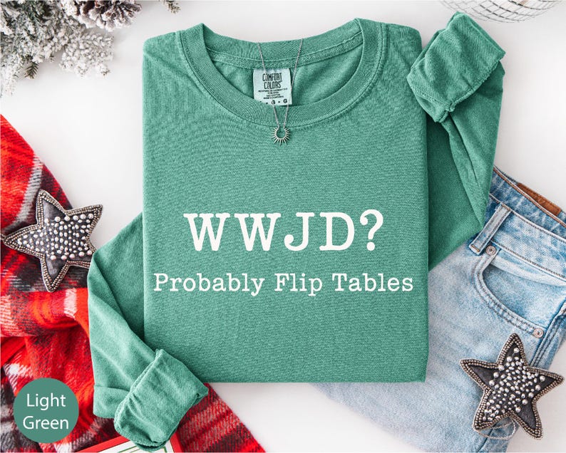 a green long-sleeved shirt with the text "WWJD?" printed on it, along with the phrase "Probably Flip Tables" below it. The shirt is laid out on a white surface, accompanied by a pair of jeans and a red plaid blanket. There are also some decorative star-shaped embellishments on the clothing and the blanket.