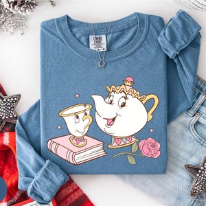 A blue t-shirt with a cartoon illustration of a teapot and a cup, along with a necklace and a pair of jeans, are displayed on a white surface.