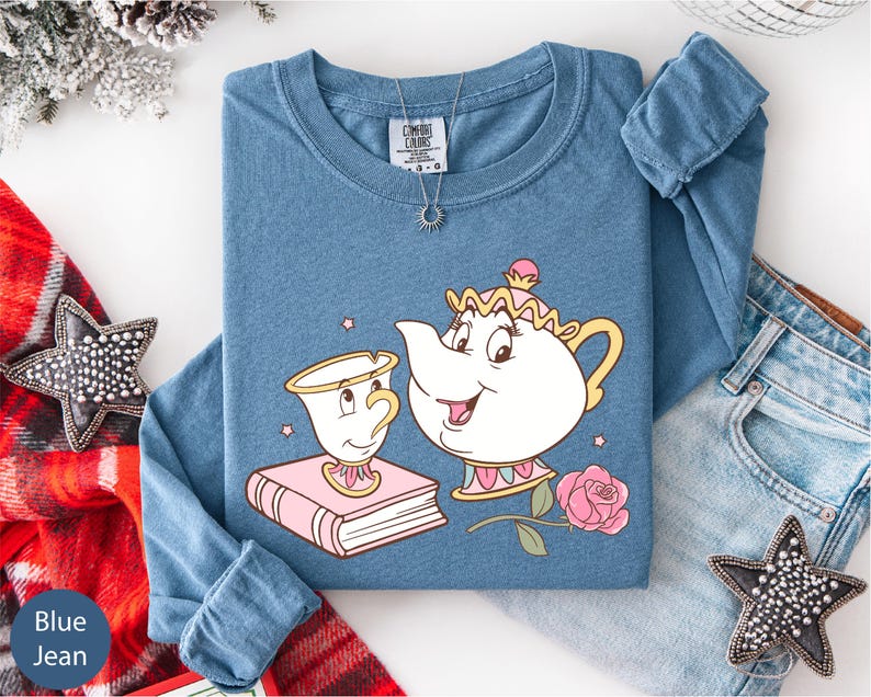 A blue t-shirt with a cartoon illustration of a teapot and a cup, along with a necklace and a pair of jeans, are displayed on a white surface.