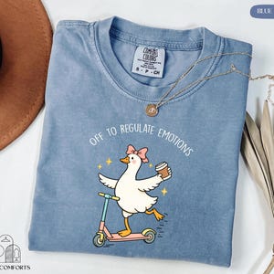 A blue t-shirt with a cartoon duck riding a scooter and holding a cup, accompanied by the text "Off to regulate emotions".