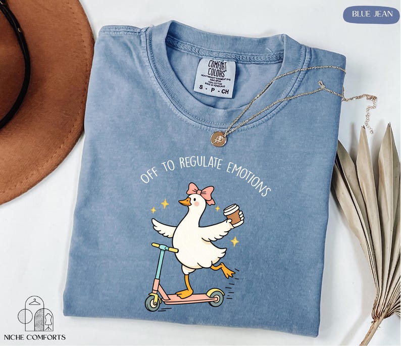 A blue t-shirt with a cartoon duck riding a scooter and holding a cup, accompanied by the text "Off to regulate emotions".