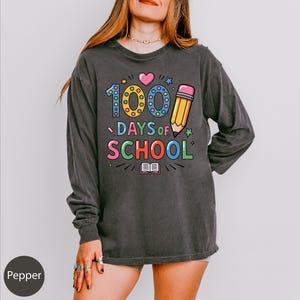 A woman wearing a gray long-sleeved shirt with a colorful graphic design that says "100 DAYS OF SCHOOL" and features a pencil, book, and heart.