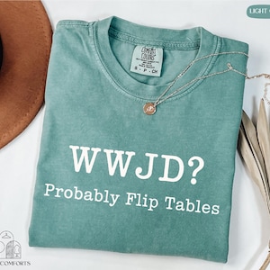 A turquoise t-shirt with the text "WWJD?" printed on it, along with the phrase "Probably Flip Tables" below it. The t-shirt is laid out on a white surface, with a brown hat and some dried leaves nearby.