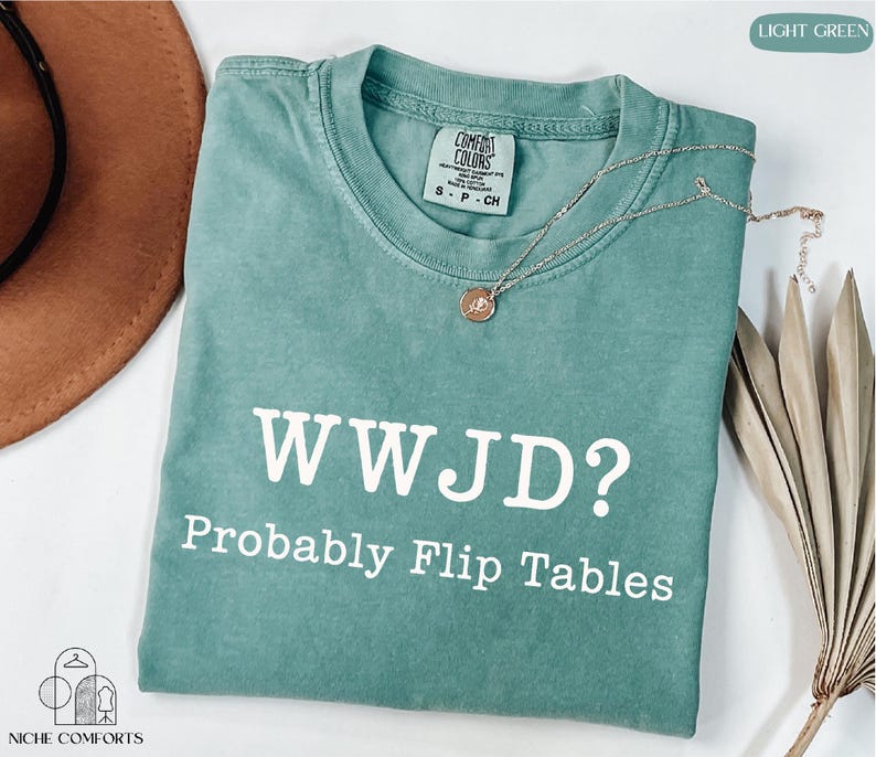 A turquoise t-shirt with the text "WWJD?" printed on it, along with the phrase "Probably Flip Tables" below it. The t-shirt is laid out on a white surface, with a brown hat and some dried leaves nearby.