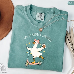 A turquoise t-shirt with a cartoon duck riding a scooter and holding a cup, accompanied by a necklace and a hat.
