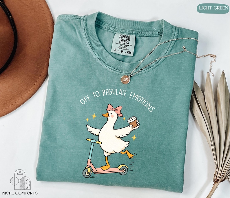 A turquoise t-shirt with a cartoon duck riding a scooter and holding a cup, accompanied by a necklace and a hat.
