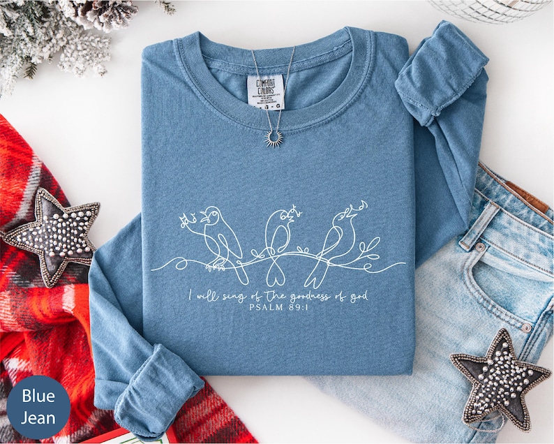 A blue t-shirt with a white design and text, along with a pair of jeans and decorative star-shaped embellishments, are arranged on a white surface.
