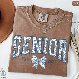 a brown t-shirt with the text "SENIOR EST. 2027" printed on it, along with a blue floral design. The t-shirt is laid out on a white surface, and there is a brown hat and some dried leaves nearby.