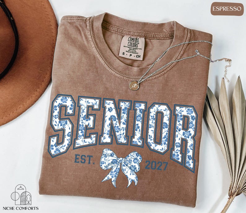 a brown t-shirt with the text "SENIOR EST. 2027" printed on it, along with a blue floral design. The t-shirt is laid out on a white surface, and there is a brown hat and some dried leaves nearby.