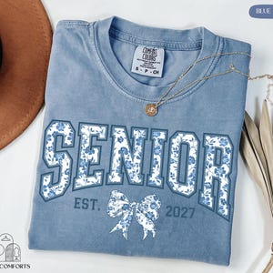 A blue t-shirt with the word "SENIOR" printed on it, along with the text "EST. 2027" below it. The t-shirt is laid out on a white surface, and there is a brown hat and some dried leaves nearby.