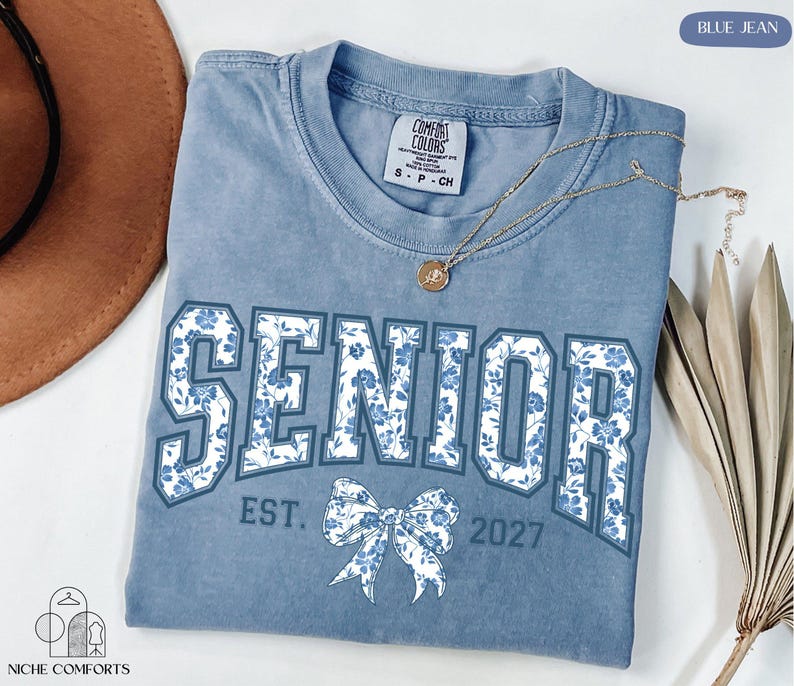 A blue t-shirt with the word "SENIOR" printed on it, along with the text "EST. 2027" below it. The t-shirt is laid out on a white surface, and there is a brown hat and some dried leaves nearby.
