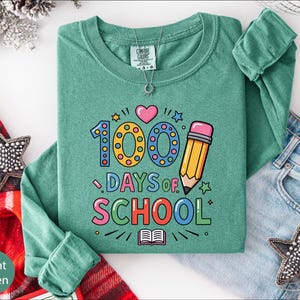 A green long-sleeved shirt with a colorful graphic design that says "100 DAYS OF SCHOOL" printed on it, along with a pencil and other school-related icons. The shirt is laid out on a white surface, with a pair of jeans and a red and white striped blanket visible in the background.