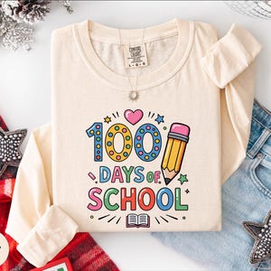 A white long-sleeved t-shirt with a colorful graphic design that says "100 DAYS OF SCHOOL" printed on it, along with various school-related icons such as a pencil, book, and star. The t-shirt is laid out on a white surface, surrounded by other clothing items like jeans and a scarf.