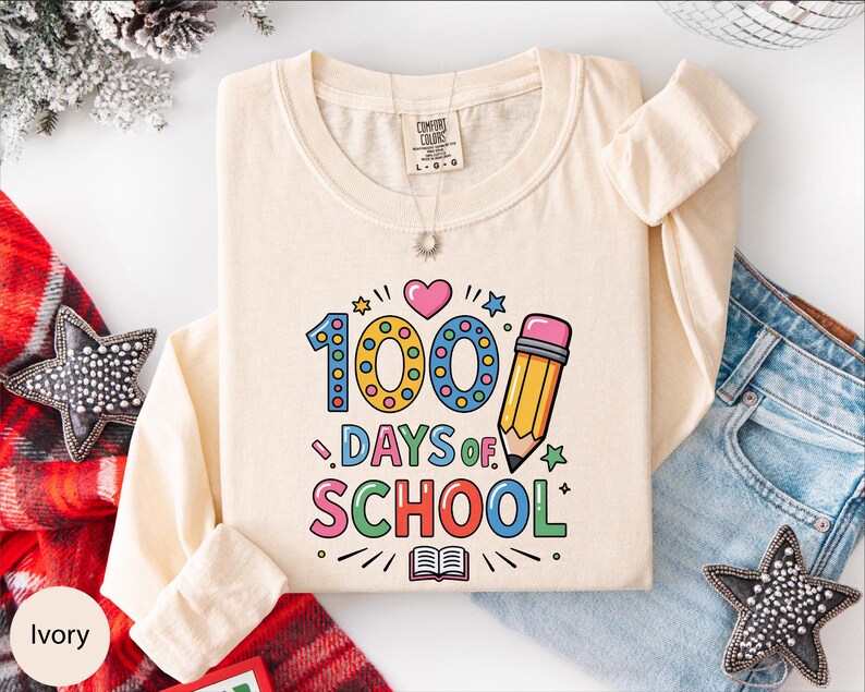 A white long-sleeved t-shirt with a colorful graphic design that says "100 DAYS OF SCHOOL" printed on it, along with various school-related icons such as a pencil, book, and star. The t-shirt is laid out on a white surface, surrounded by other clothing items like jeans and a scarf.