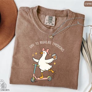 a brown t-shirt with a cartoon image of a duck riding a scooter, accompanied by the text "Off to regulate emotions".