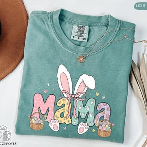 May include: Light green t-shirt with a colorful Easter-themed graphic. The design features the word "Mama" with bunny ears, eggs, and baskets. A silver necklace with a pendant is draped across the shirt. The shirt is labeled "Comfort Colors".