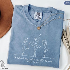 May include: Blue jean Comfort Colors t-shirt with a white line drawing of three ballet dancers. The shirt features the text "He turned my mourning into dancing. Psalm 30:11-12." A gold necklace rests on the shirt.
